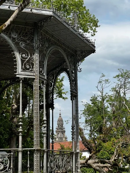 Quiosco de música de hierro forjado con ornamentación modernista en la Alameda de Santiago de Compostela, con la torre del reloj de la Catedral visible al fondo entre los árboles.