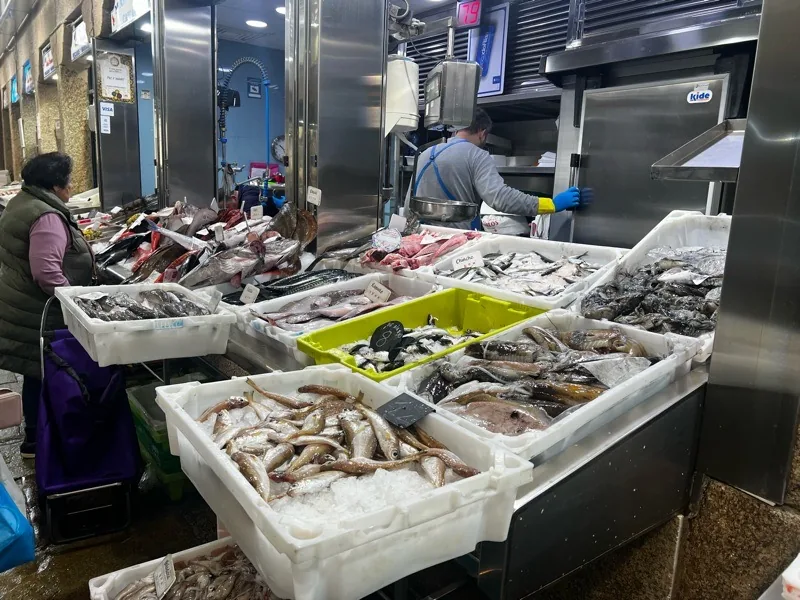 Puesto de pescado fresco en el Mercado de Abastos de Santiago de Compostela, con cajas de plástico blanco y verde llenas de diversas especies de pescado sobre hielo, un vendedor trabajando al fondo y una clienta a la izquierda.