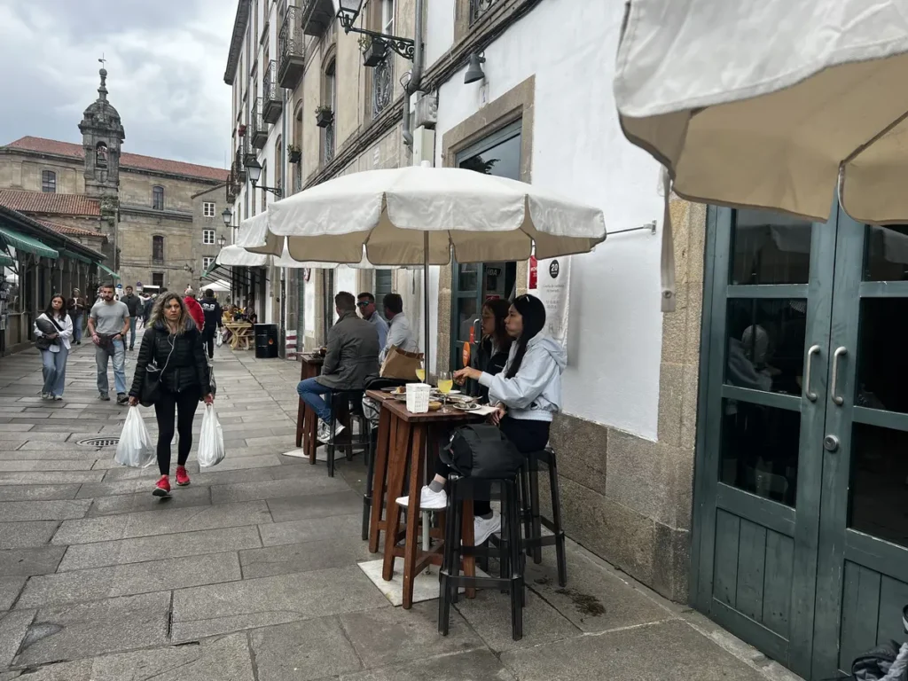 Terraza exterior del restaurante Abastos 2.0 junto al Mercado de Abastos de Santiago de Compostela, con barra corrida bajo toldos verdes y mesas altas en la calle empedrada del casco histórico.
