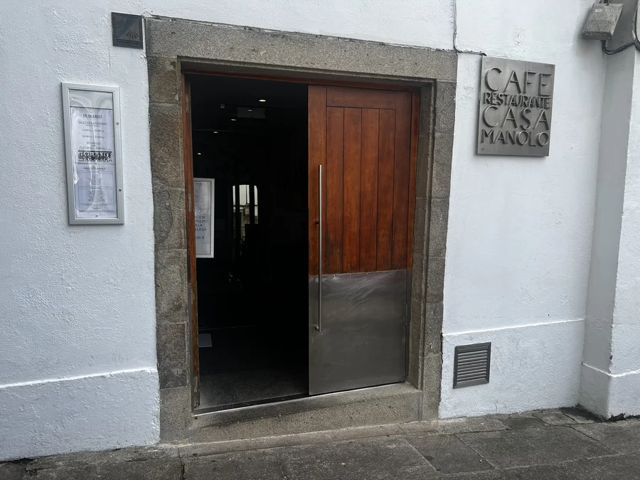 Fachada, interior y menú del día del Café Restaurante Casa Manolo en Santiago de Compostela: entrada con puerta de madera y cartel en piedra gallega, sala interior con decoración tradicional gallega y menú de dos platos por 14,50 € con caldo gallego, croquetas caseras, pescados y carnes a la plancha.