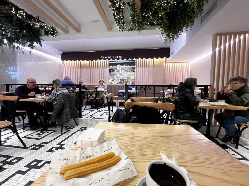 Interior de la Churrería La Quinta en Santiago de Compostela, con churros recién hechos y chocolate a la taza sobre una mesa de madera, en un local de diseño moderno.