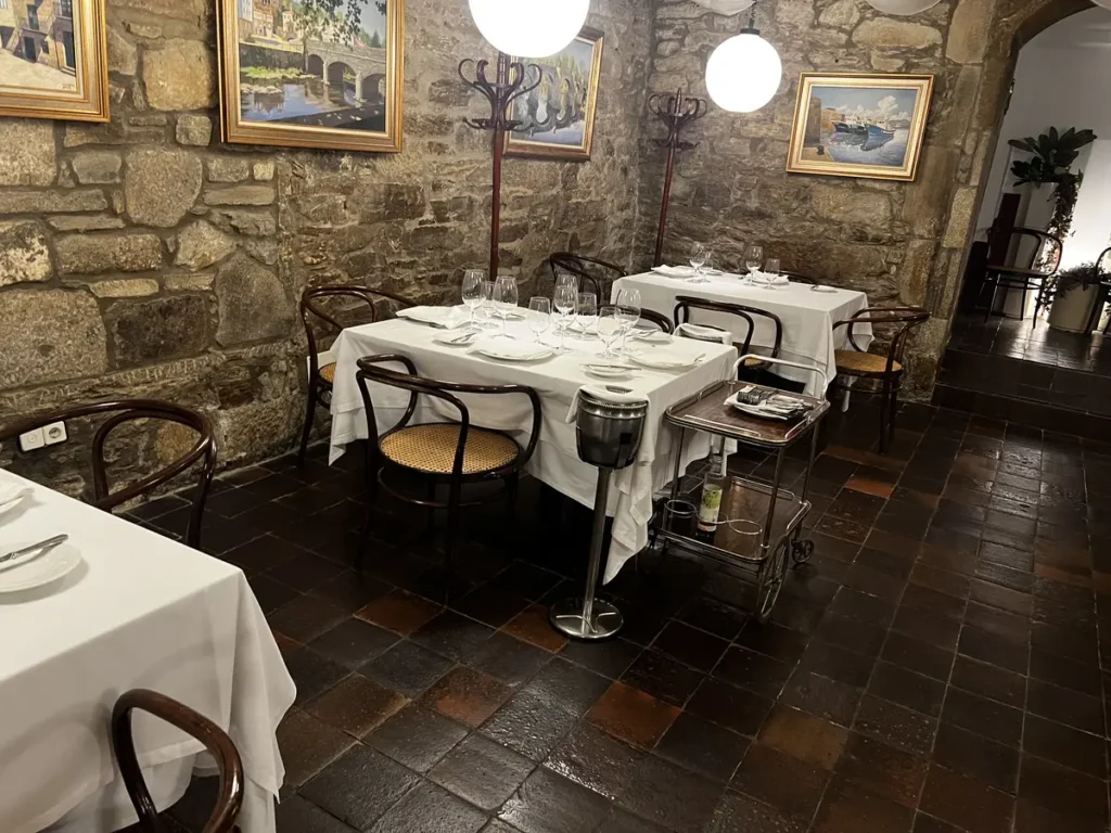 Interior del Restaurante Don Gaiferos en Santiago de Compostela: salón con bóvedas de piedra centenaria, mesas con manteles blancos y cristalería, decoración con pinturas gallegas, y plato de pulpo a feira con pimentón.