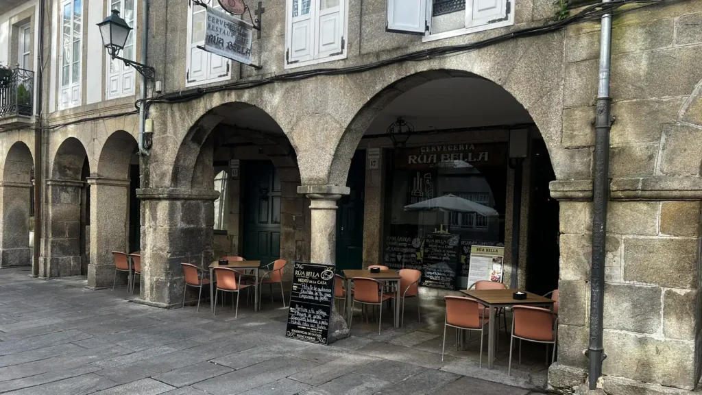 Terraza de la Cervecería Rúa Bella bajo los soportales de piedra del casco histórico de Santiago de Compostela, con mesas en la calle empedrada protegidas de la lluvia