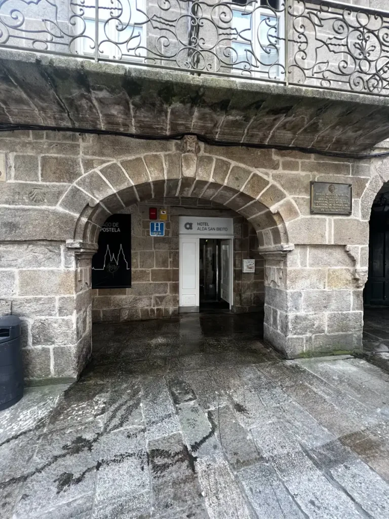 Fachada, entrada y placa histórica del Hotel Alda San Bieito en Santiago de Compostela: edificio de piedra centenaria con balcones de forja, acceso por arco de cantería y placa que recuerda que en este edificio vivió Manuel Martínez Murguía, insigne precursor del galleguismo.