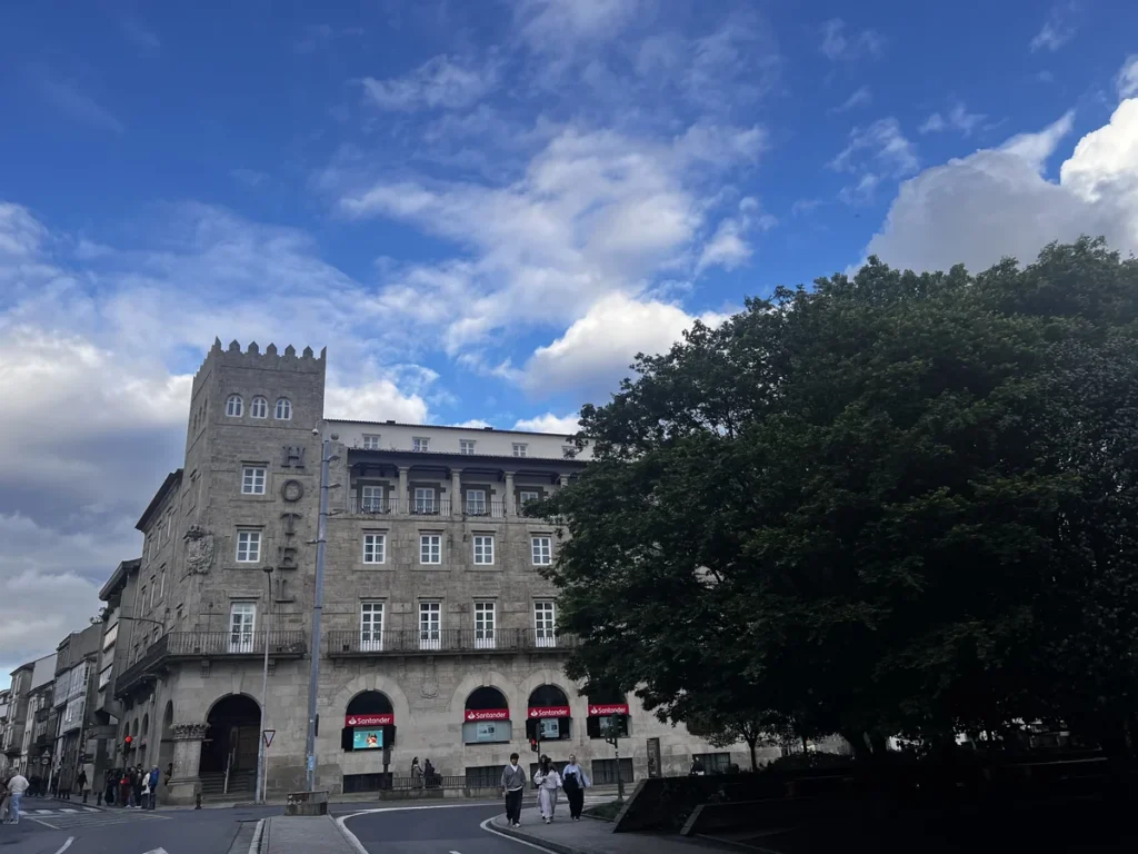 Fachada de piedra del Hotel Compostela en Santiago de Compostela con cielo azul y gente paseando​​​​​​​​​​​​​​​​ por la siempre transitada plaza de Galicia