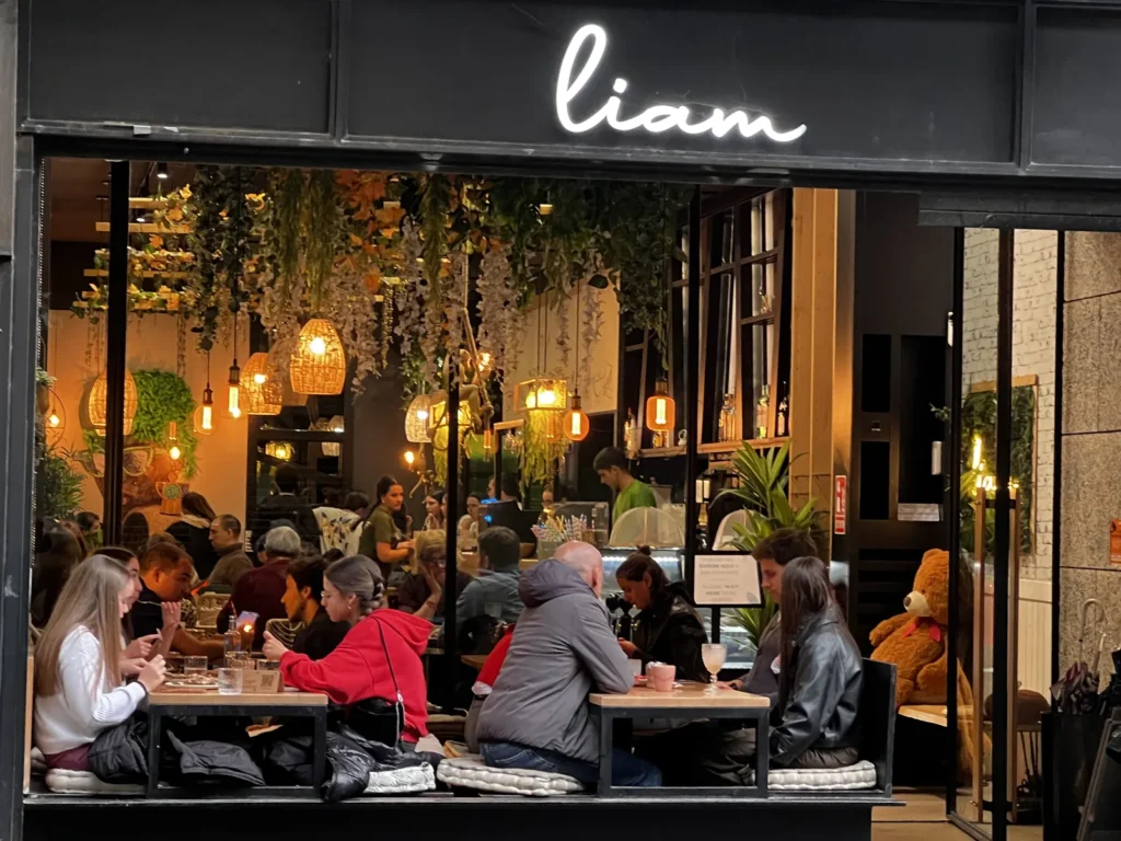 Terraza e interior del café boutique Liam en Santiago de Compostela, con mesas llenas de clientes disfrutando del ambiente, iluminado con lámparas cálidas y vegetación colgante.