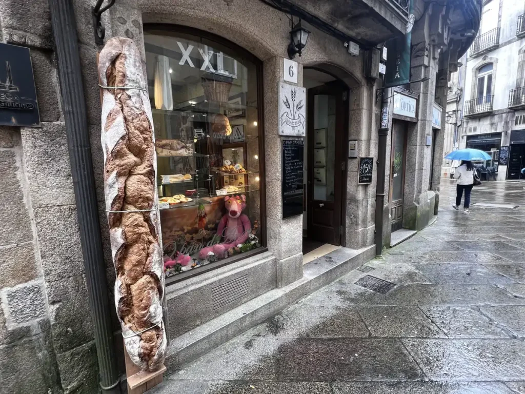 Fachada de la panadería Pan d'Andrea en Santiago de Compostela, con una enorme barra de pan artesano decorativa en la columna de piedra y escaparate con empanadas y bollería