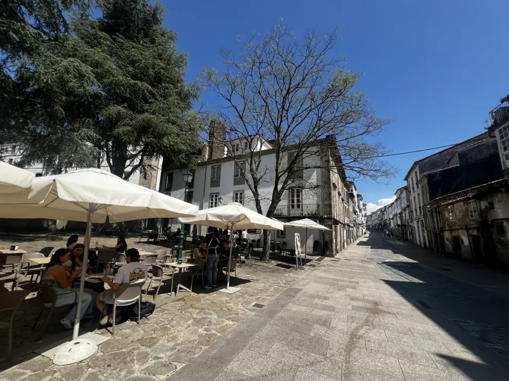Rúa de San Pedro en Santiago de Compostela: calle empedrada con las torres de la catedral al fondo y terrazas con sombrillas blancas, donde conviven restaurantes como A Maceta, De Lito, O Dezaseis y el premiado PAMPÍN Bar.